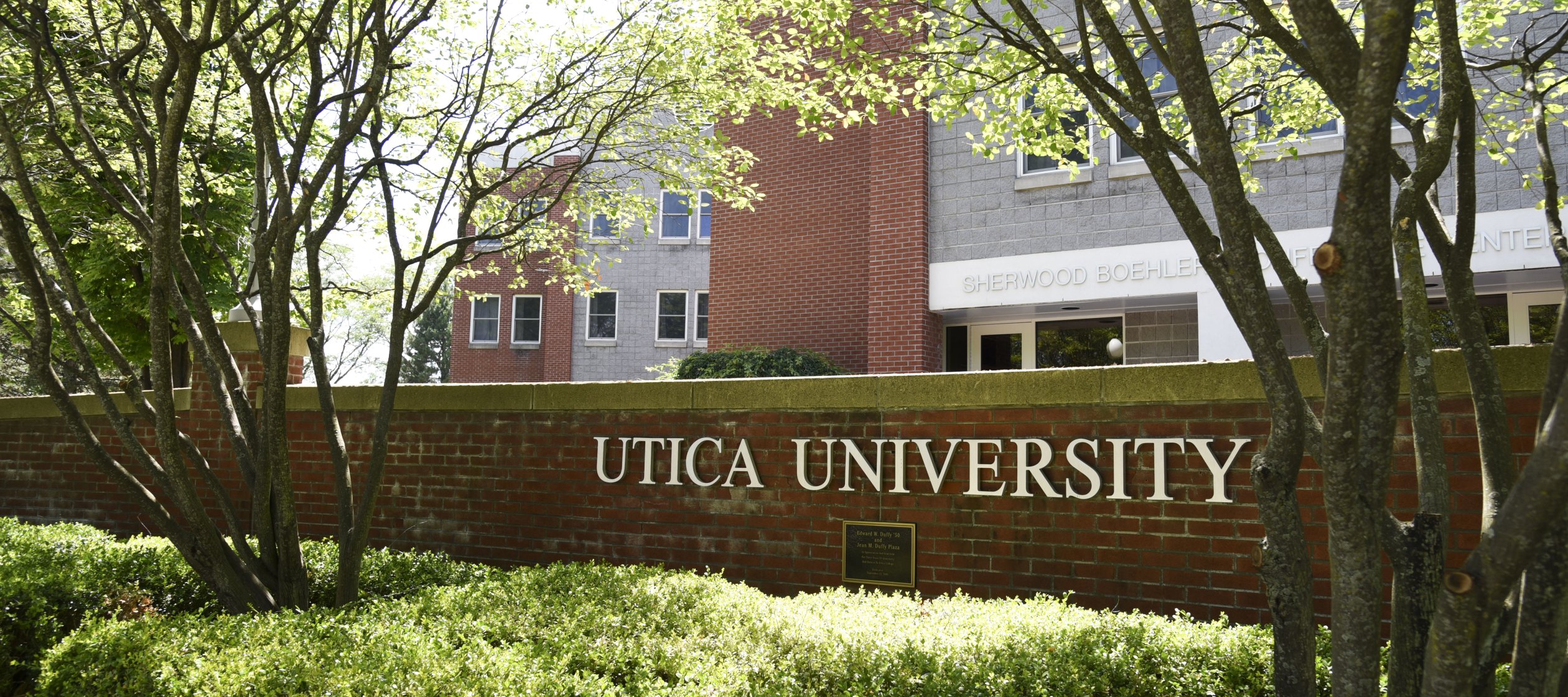 The words Utica University against a brick wall in front of a residence hall.