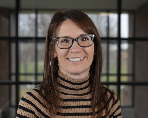 Renee Fisher in glasses and a striped turtleneck stands in front of a window.